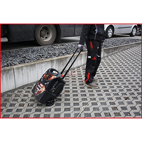 Person zieht eine schwarze Trolley-Box mit Rädern über ein Pflaster. Im Hintergrund ein Bus und ein Auto.